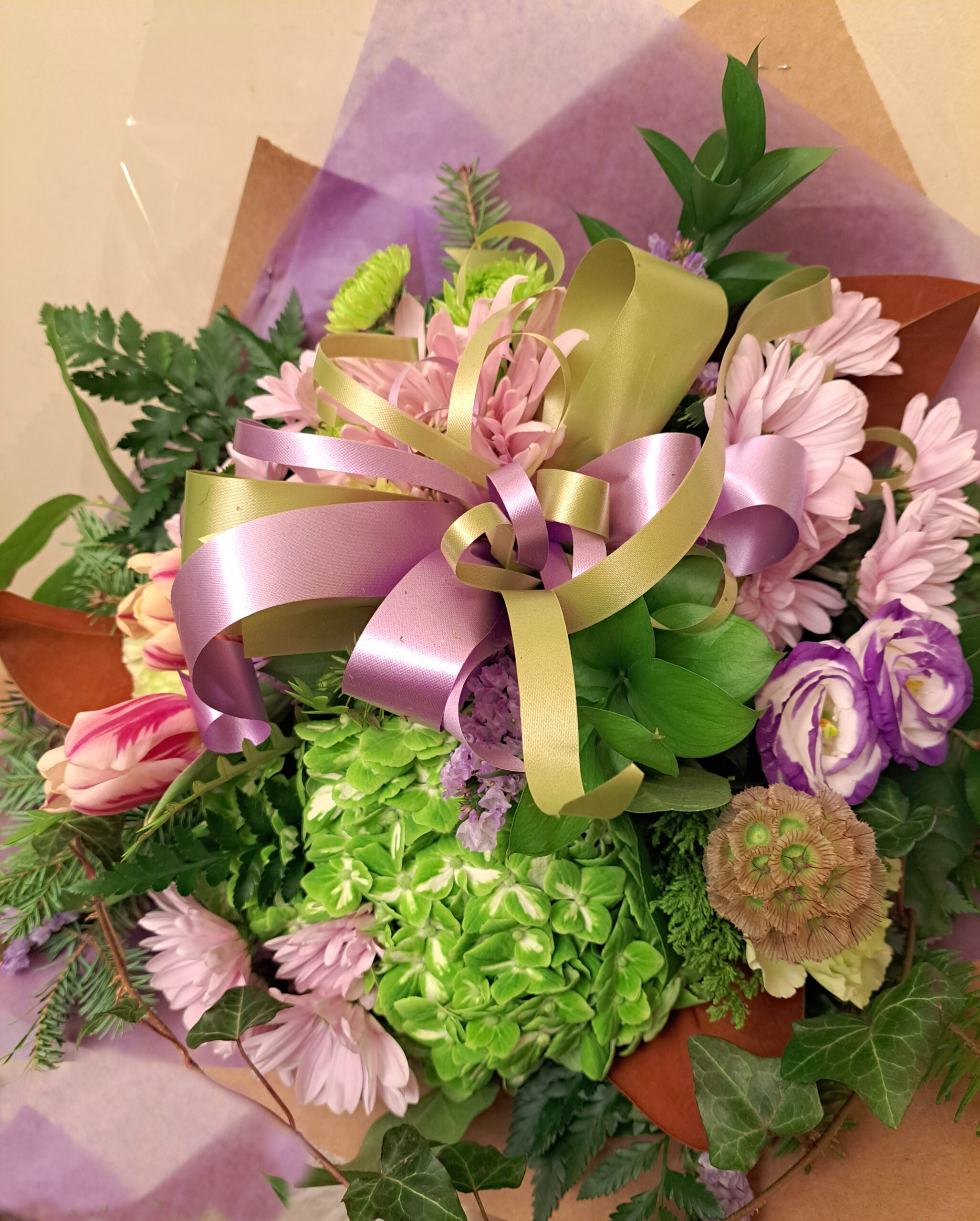 Bouquet of pink flowers, green foliage, and pastel ribbons tied with gold ribbon, wrapped in purple paper.