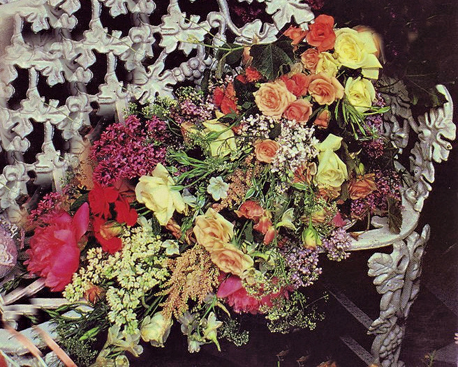 Large colorful funeral bouquet with roses and mixed flowers resting on an ornate white chair or bier.