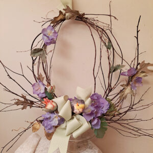 Decorative twig wreath with purple flowers and a pale ribbon against a beige wall surface.