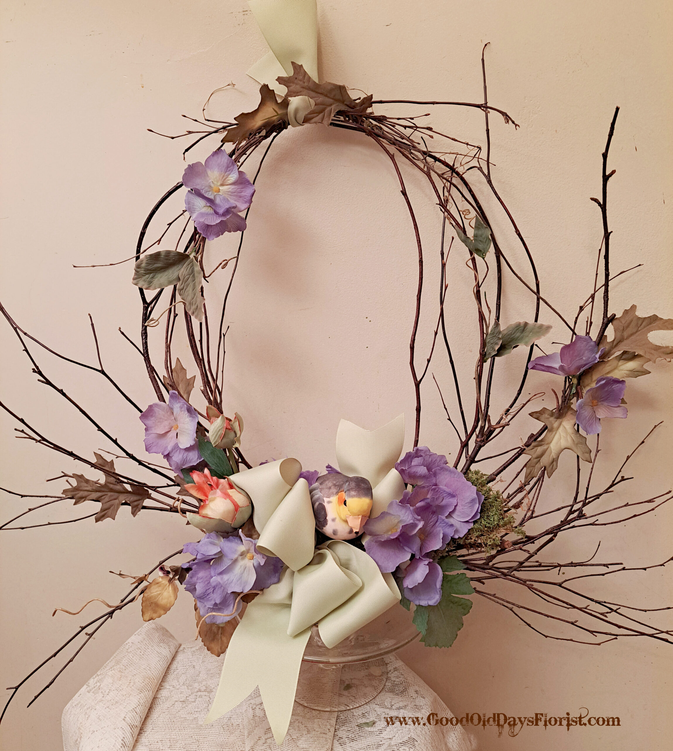 Decorative twig wreath with purple flowers and a pale ribbon against a beige wall surface.