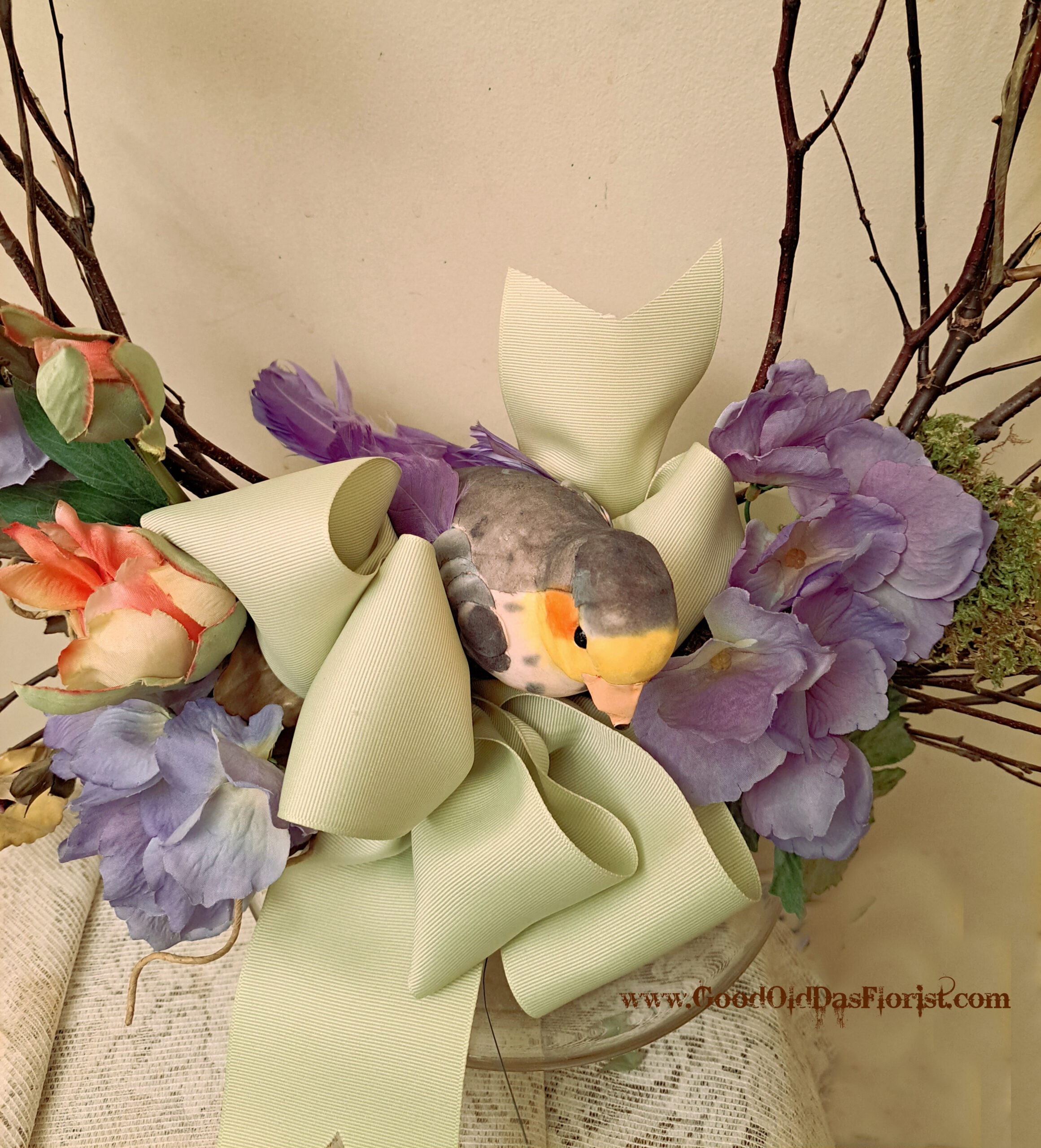 Close-up of a floral arrangement with purple hydrangeas, pale green ribbons, and twisting branches against a light background (watermark visible).