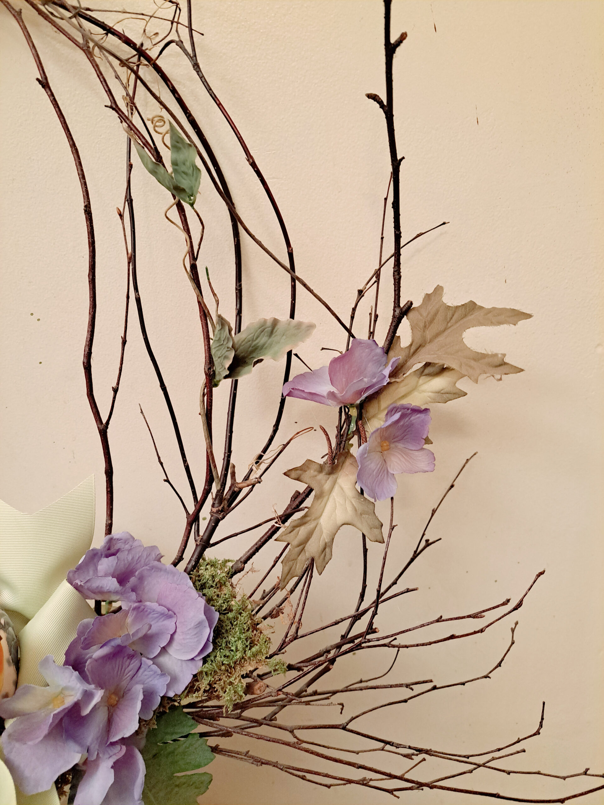 Decorative floral arrangement featuring purple blossoms and dried brown branches against a neutral wall, with mossy base and pale ribbon.