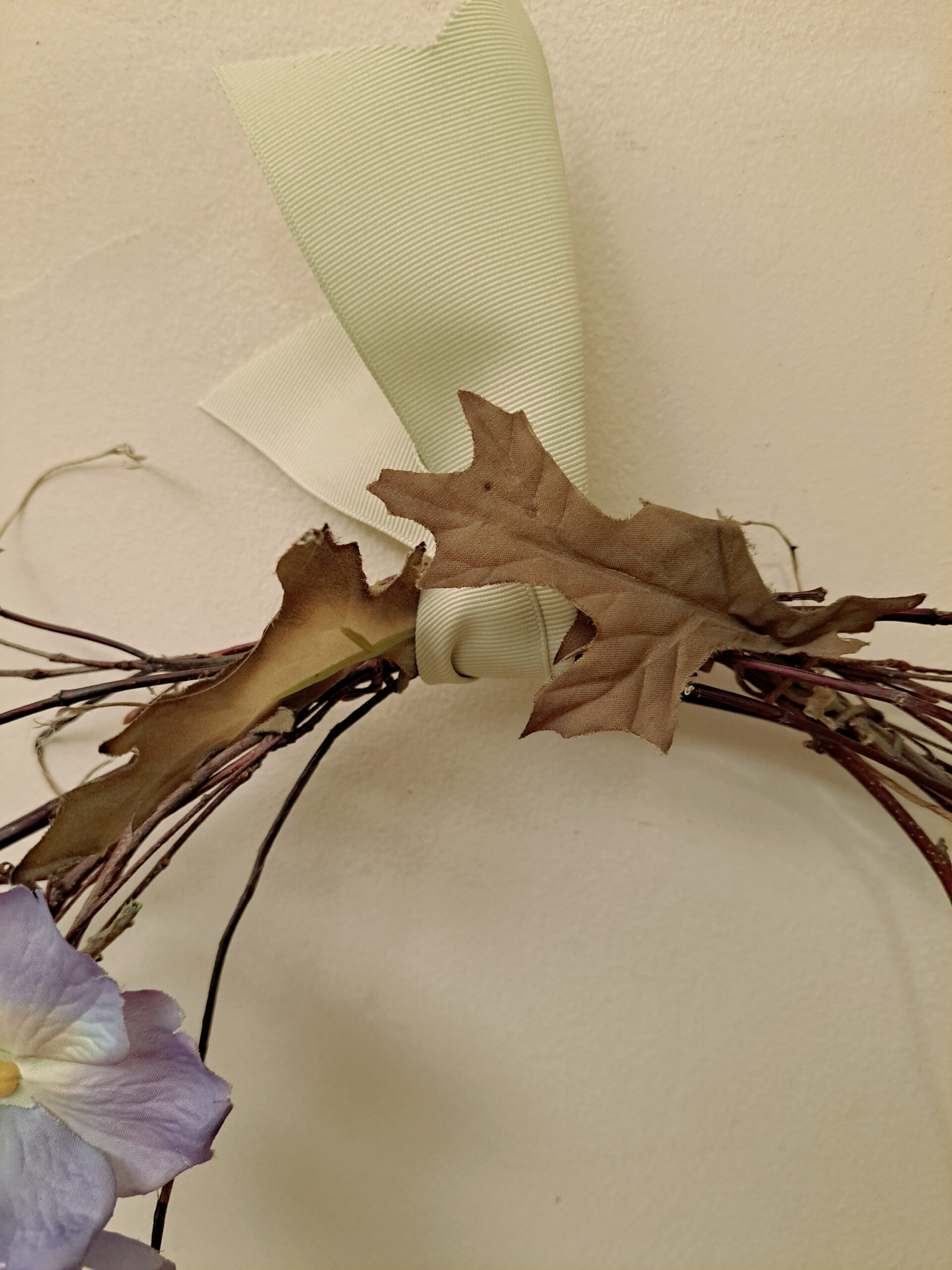 Close-up of a wreath with brown dried leaves, a pale green ribbon, and a lavender fabric flower.