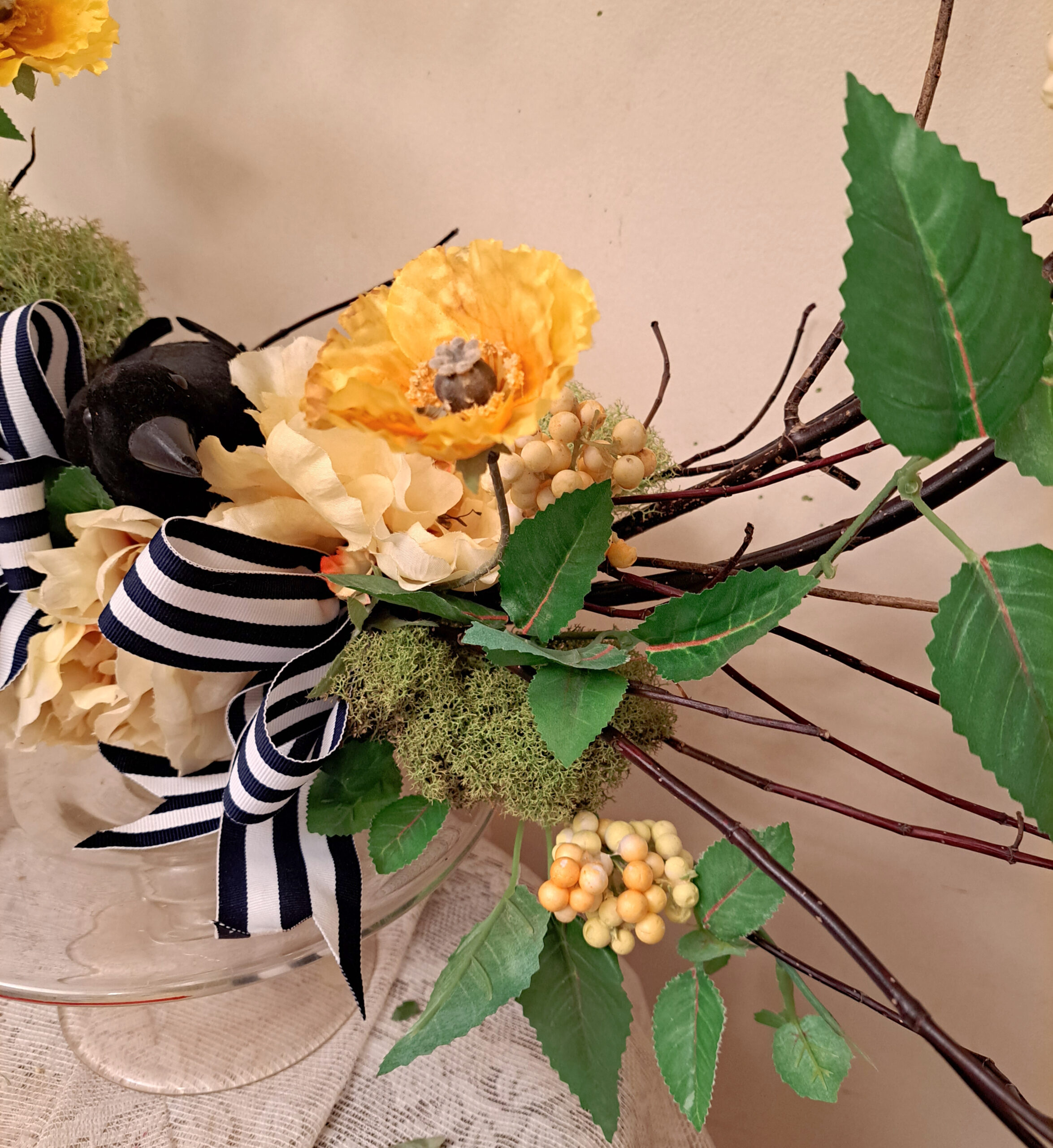 Decorative floral arrangement with pale yellow flowers, greenery, moss, and a black-and-white striped ribbon in a glass vase.
