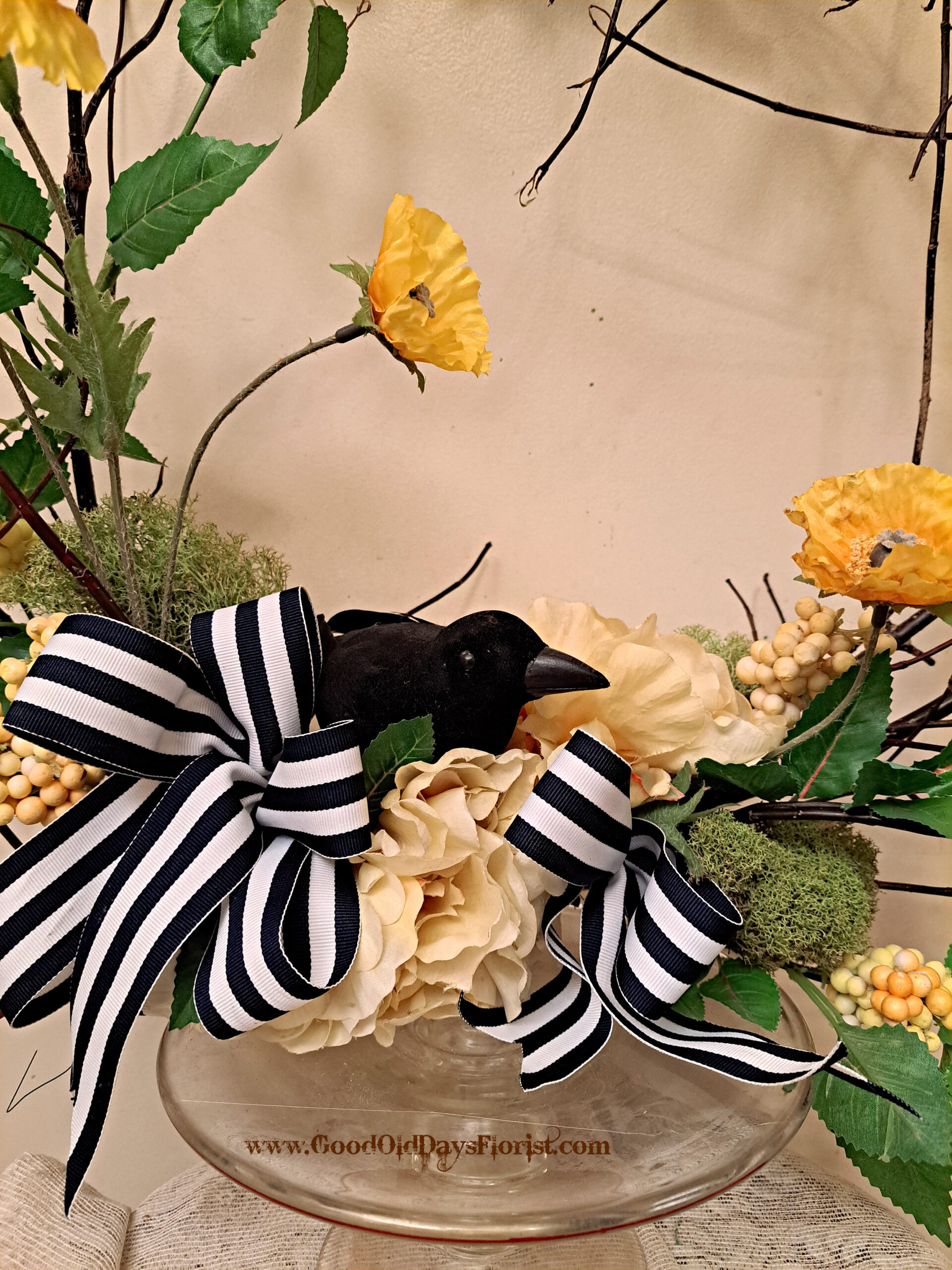 Decorative floral centerpiece with cream and yellow flowers, green leaves, a black bird figure, and a bold black-and-white striped ribbon in a glass vase.